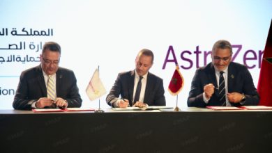 Three men in suits sign documents at a formal ceremony, with small flags on the table and a backdrop showing Arabic text and the AstraZeneca logo.