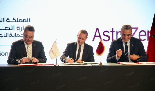 Three men in suits sign documents at a formal ceremony, with small flags on the table and a backdrop showing Arabic text and the AstraZeneca logo.