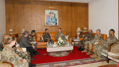 Military officers in camouflage uniforms sit in a formal briefing room around a central coffee table with a flower arrangement, wood-paneled wall backdrop, and a portrait above.