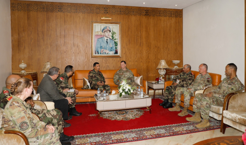 Military officers in camouflage uniforms sit in a formal briefing room around a central coffee table with a flower arrangement, wood-paneled wall backdrop, and a portrait above.