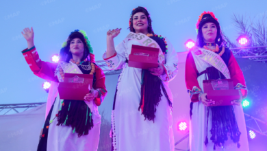 Three women on a stage wearing sashes and white dresses, holding pink boxes as colorful stage lights illuminate them (pageant or festival setting).