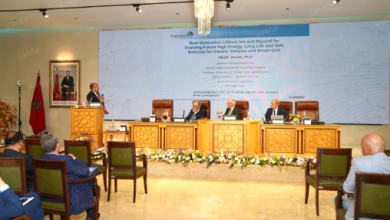 Speaker at a podium addresses a panel discussion on a conference stage with a large screen, Moroccan flag, and an ornate wooden dais.