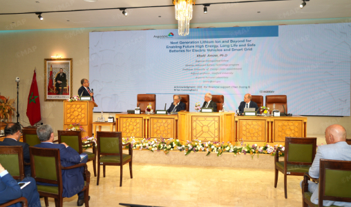 Speaker at a podium addresses a panel discussion on a conference stage with a large screen, Moroccan flag, and an ornate wooden dais.
