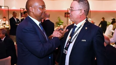 Two men in dark suits at a formal event converse, one adjusting the other’s lapel with a FIFA badge visible, guests and a buffet in the background.