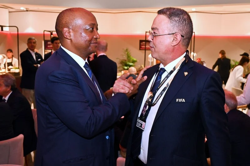 Two men in dark suits at a formal event converse, one adjusting the other’s lapel with a FIFA badge visible, guests and a buffet in the background.