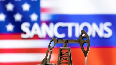 Miniature oil drilling rig in the foreground with a blurred US flag and the word SANCTIONS in the background.