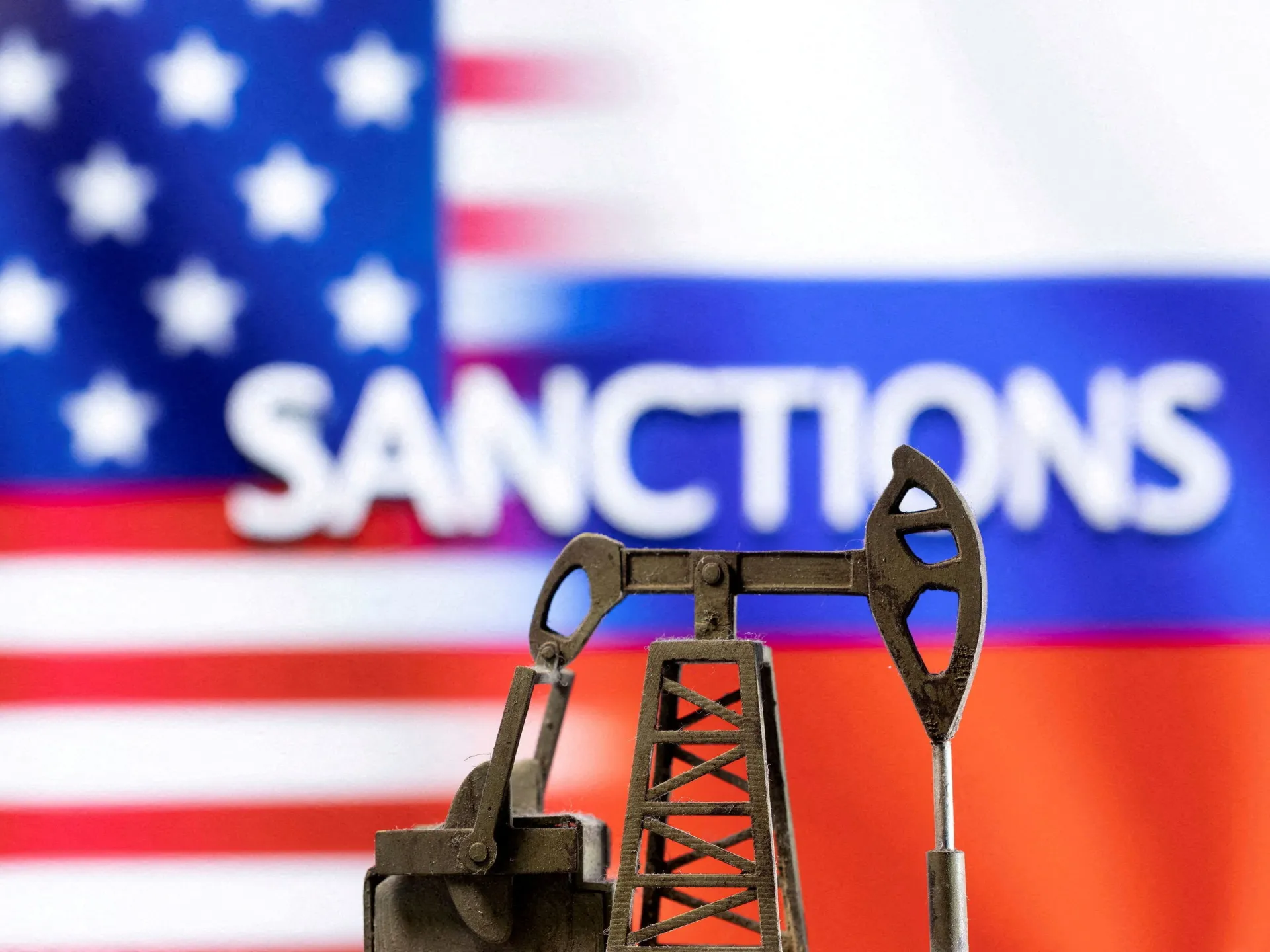 Miniature oil drilling rig in the foreground with a blurred US flag and the word SANCTIONS in the background.