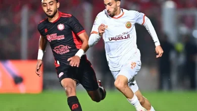 Two soccer players sprint side by side on a green pitch, chasing the ball during a match under stadium lights.
