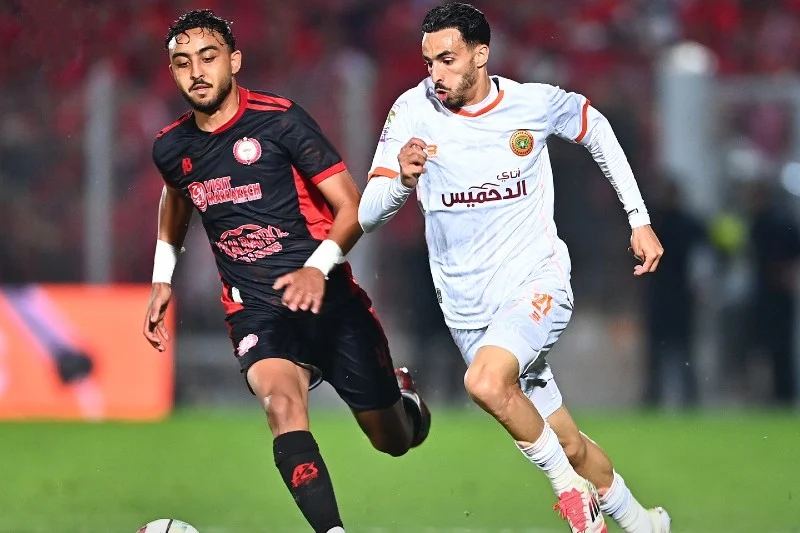 Two soccer players sprint side by side on a green pitch, chasing the ball during a match under stadium lights.