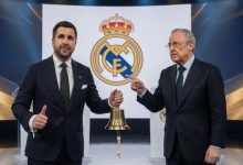 Two men in dark suits stand on a stage, one holding a brass bell as they pose with a Real Madrid crest backdrop behind them.