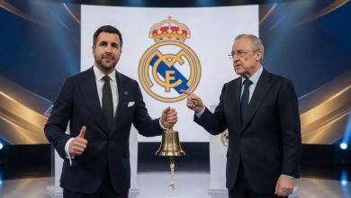 Real-Madridjpg | MCG24 | موقع إلكتروني مغربي شامل متجدد على مدار الساعة Two men in dark suits stand on a stage, one holding a brass bell as they pose with a Real Madrid crest backdrop behind them.