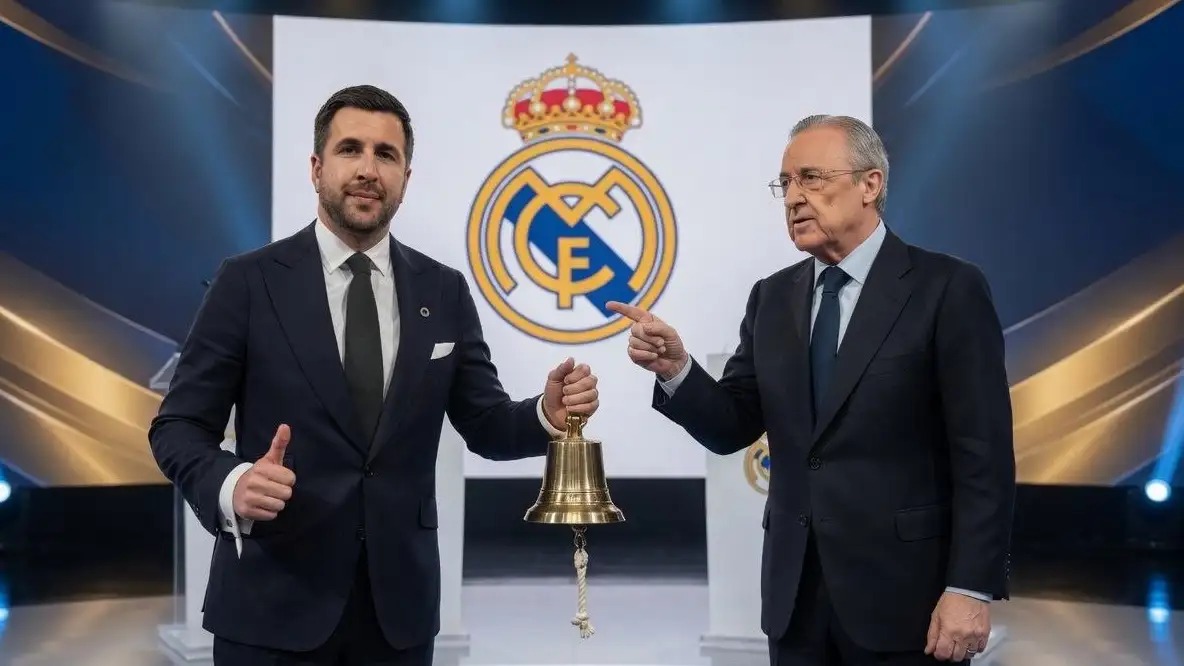 Real-Madridjpg | MCG24 | موقع إلكتروني مغربي شامل متجدد على مدار الساعة Two men in dark suits stand on a stage, one holding a brass bell as they pose with a Real Madrid crest backdrop behind them.