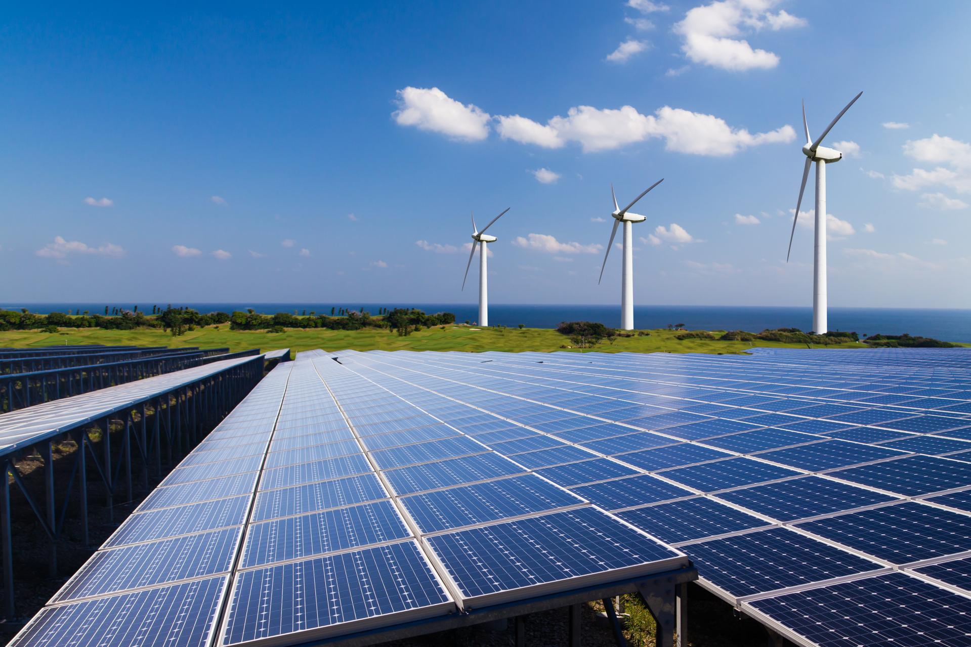 Rows of solar panels in the foreground with three wind turbines on a grassy hill near the coast under a clear blue sky.