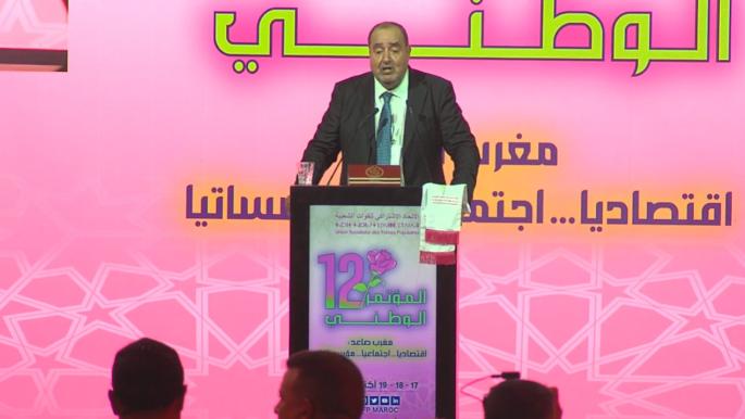 Screenshot 2025-10-18 040647 | MCG24 | موقع إلكتروني مغربي شامل متجدد على مدار الساعة Man in a suit giving a speech at a podium on a bright pink conference stage with Arabic signage in the background.