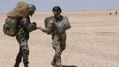 Two soldiers in desert camouflage shake hands, one carrying a large pack, under a clear blue sky, as they meet on flat sand terrain.