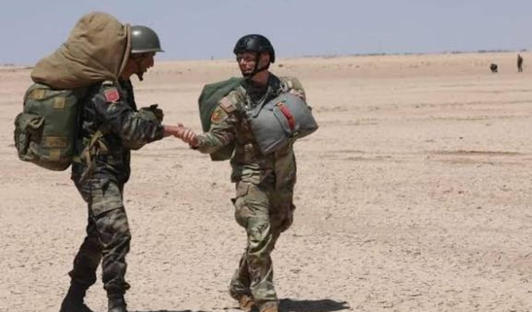 Two soldiers in desert camouflage shake hands, one carrying a large pack, under a clear blue sky, as they meet on flat sand terrain.