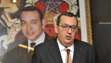 Man in a dark pinstripe suit and orange tie speaking into a microphone at a podium, wearing glasses with a red lapel pin, with a large framed portrait behind him.