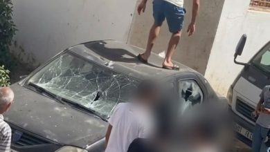 Man wearing a white t-shirt and blue shorts stands on the roof of a dark car with a shattered windshield, while several people watch nearby.