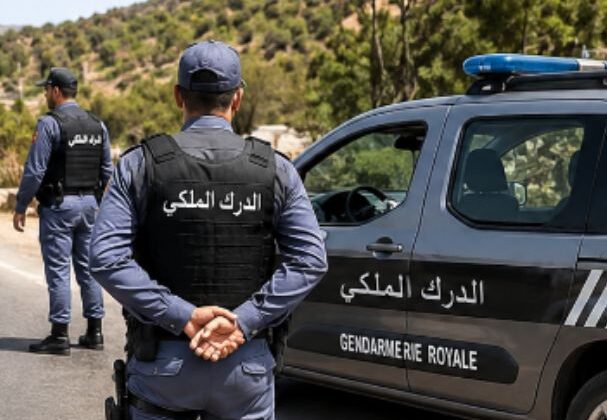 Screenshot_20260507-141724-607x420 | MCG24 | موقع إلكتروني مغربي شامل متجدد على مدار الساعة Two Royal Gendarmerie officers in blue uniforms stand beside a marked patrol car on a rural road, with Arabic labels on jackets and vehicle.