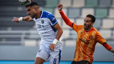 Two soccer players compete for the ball during a match, one in a white kit and the other in orange, both reaching for the ball midair.