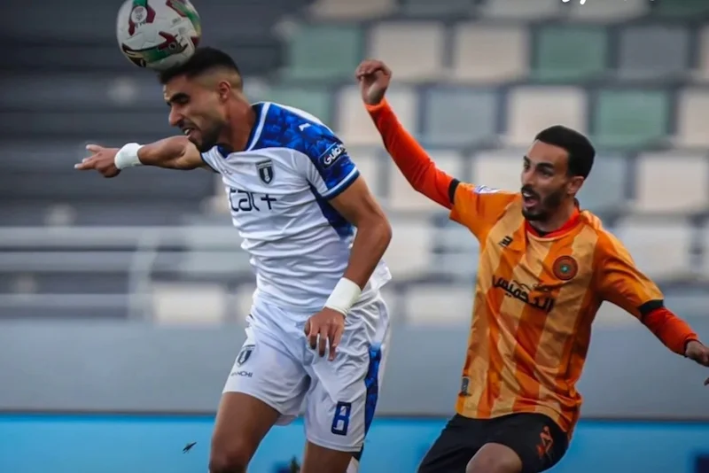 Two soccer players compete for the ball during a match, one in a white kit and the other in orange, both reaching for the ball midair.
