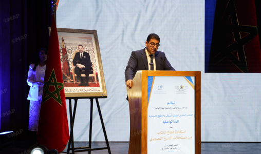Man in a suit delivering a speech at a podium on a stage, with the Moroccan flag and a framed portrait nearby.