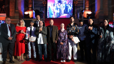 Group of diverse people on a red carpet posing for a photo at an awards event; several hold certificates with a large screen and stage lighting in the background.