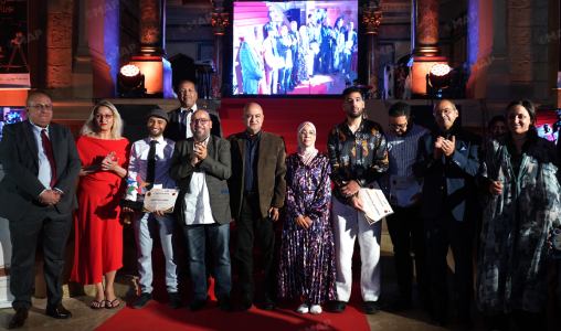 Group of diverse people on a red carpet posing for a photo at an awards event; several hold certificates with a large screen and stage lighting in the background.