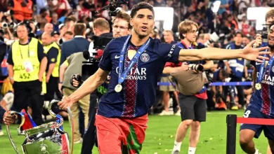 PSG footballer with medal, arms outstretched, celebrating on the field beside a large trophy. The crowd cheers in the background.