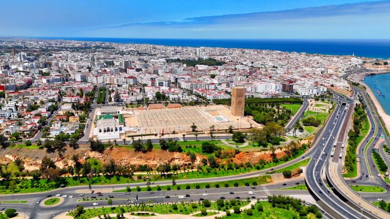 VCB7YNKOQZAUJHLATXHCFVCMLI-768x432 | MCG24 | موقع إلكتروني مغربي شامل متجدد على مدار الساعة Aerial view of a coastal city with a large square and a tall tower, surrounding streets, and a winding highway, sea in the background.