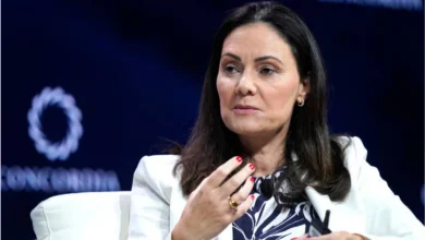 Woman with dark hair in a white blazer speaks on a panel, gesturing with her hand while seated on stage. (Note: stage backdrop is dark with blurred logos)