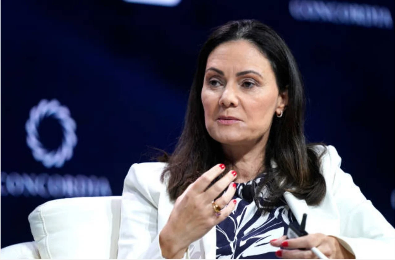 Woman with dark hair in a white blazer speaks on a panel, gesturing with her hand while seated on stage. (Note: stage backdrop is dark with blurred logos)