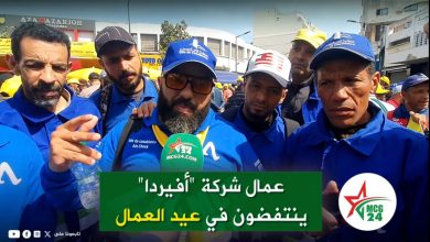 Group of men in blue uniforms being interviewed on a busy street, with a green Arabic banner and an MCG24 microphone in the foreground.