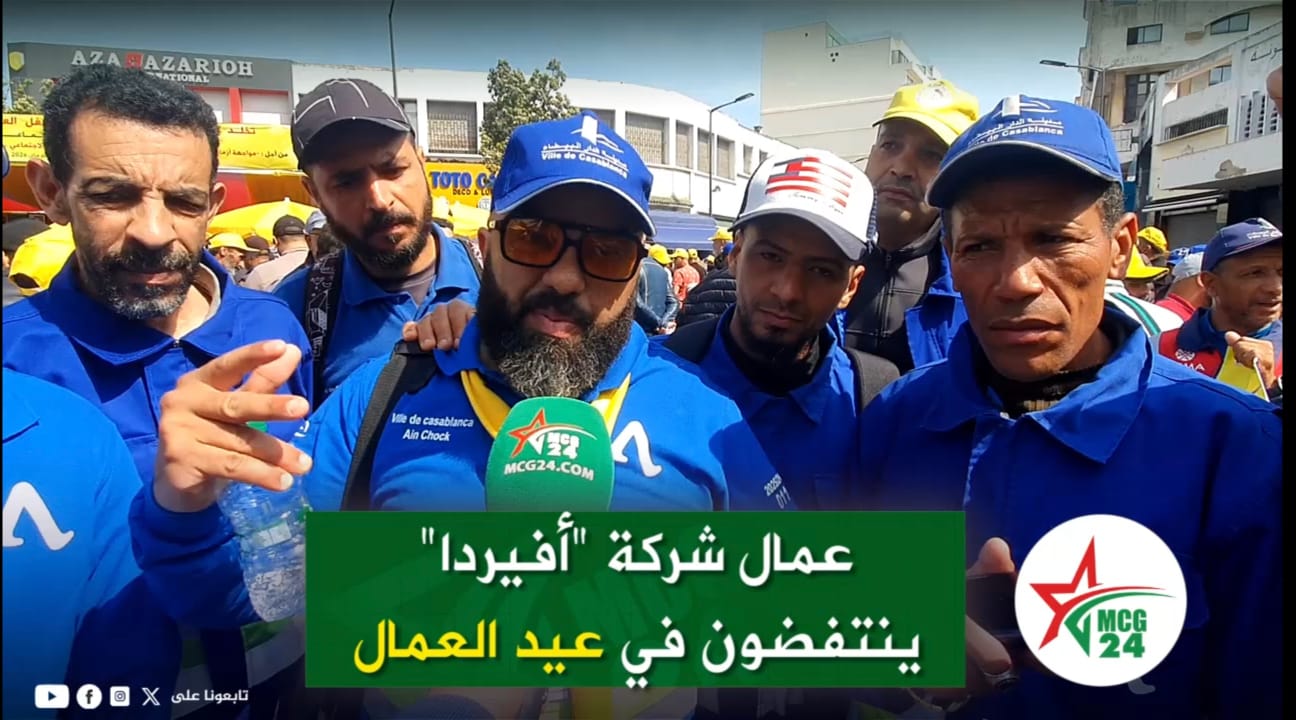 Group of men in blue uniforms being interviewed on a busy street, with a green Arabic banner and an MCG24 microphone in the foreground.