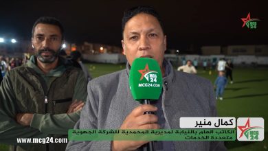 Male reporter holding a green MCG24 microphone speaks at a night-time sports field, with a teammate beside him and a crowd in the background; Arabic caption overlaid at bottom with MCG24 branding.