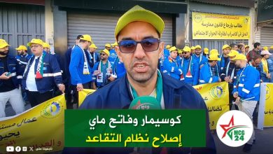 Reporter in yellow cap speaks to the camera as a crowd of protesters in blue jackets and yellow hats line a street behind him, carrying banners and signs in Arabic.
