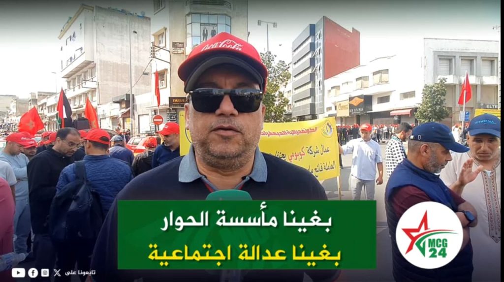 Male reporter wearing a red cap and sunglasses speaks to the camera at a street protest, with a crowd, red flags, and a yellow banner in the background; MC G24 news logo bottom right.