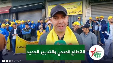 Man with a dark cap and yellow scarf speaks to the camera as a street protest unfolds behind him, banners and a crowd in blue and yellow visible in the background.