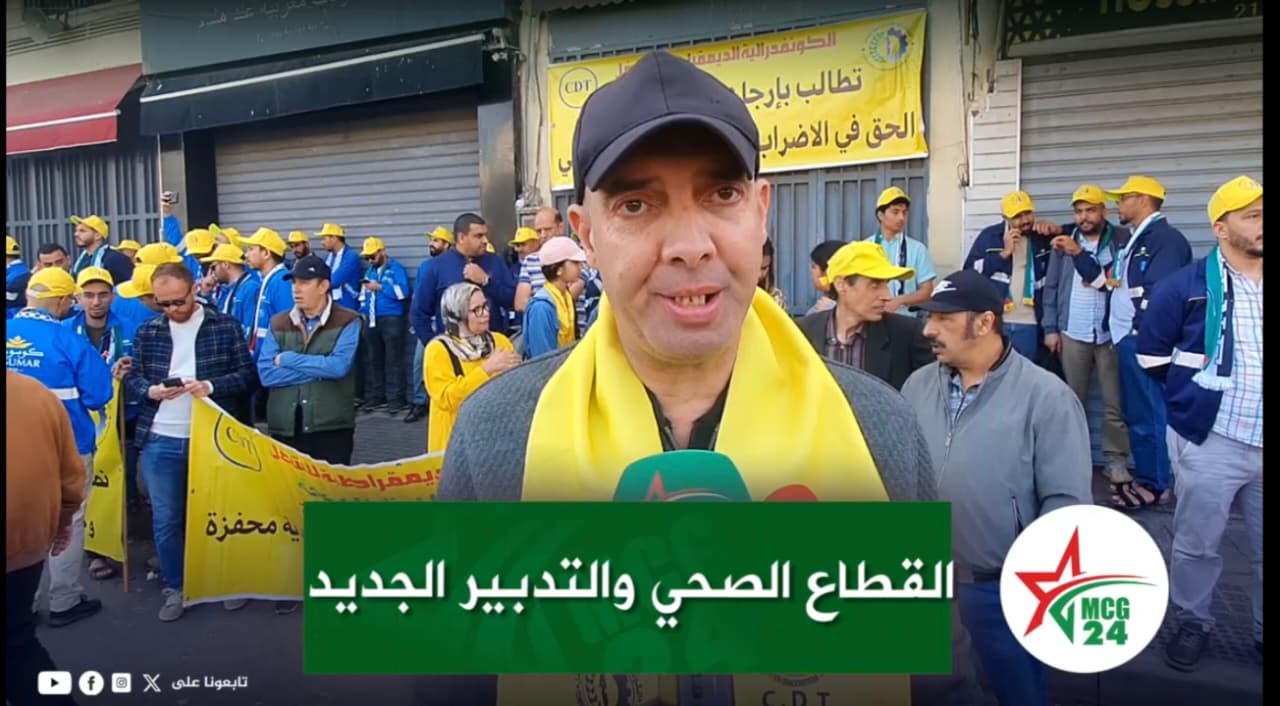 Man with a dark cap and yellow scarf speaks to the camera as a street protest unfolds behind him, banners and a crowd in blue and yellow visible in the background.