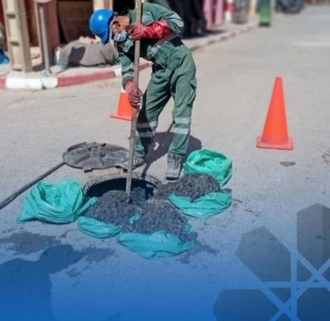 Road construction worker in green overalls and blue helmet shoveling hot asphalt into a pothole, orange traffic cones and green asphalt bags nearby.
