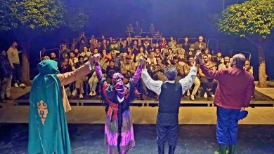 Four performers stand on a stage facing a seated audience with arms raised in a gesture, outdoors at night with trees in the background.
