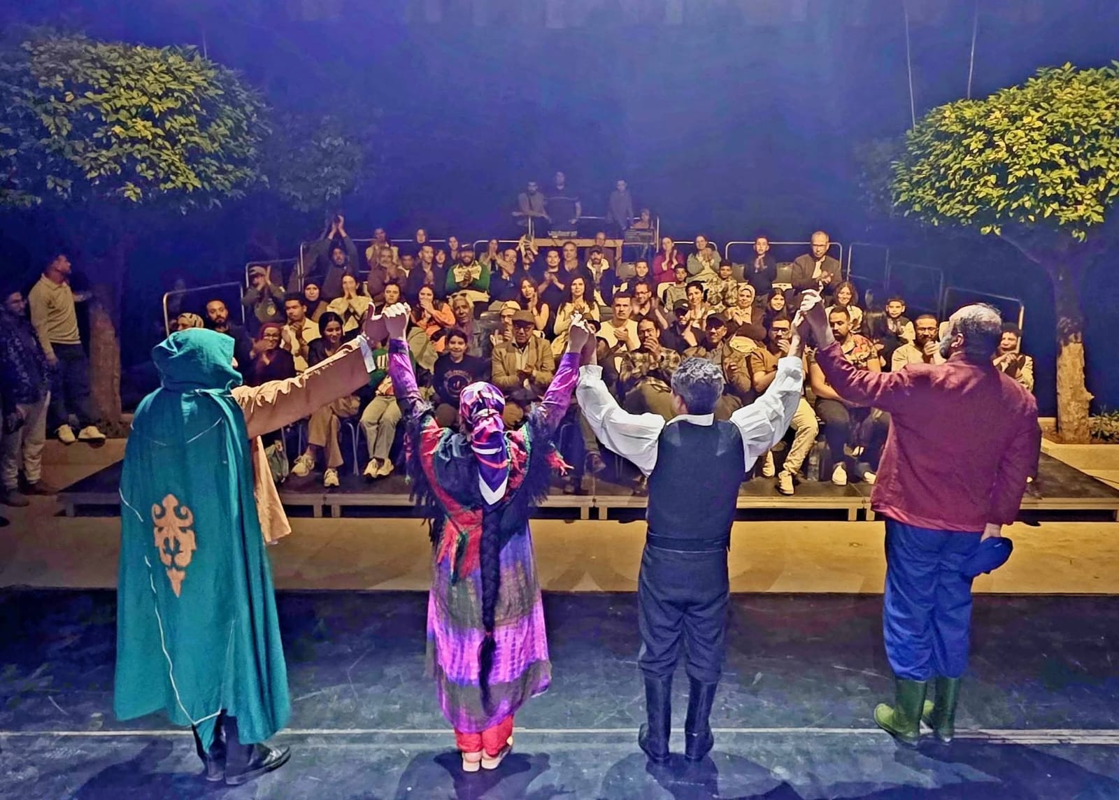 Four performers stand on a stage facing a seated audience with arms raised in a gesture, outdoors at night with trees in the background.