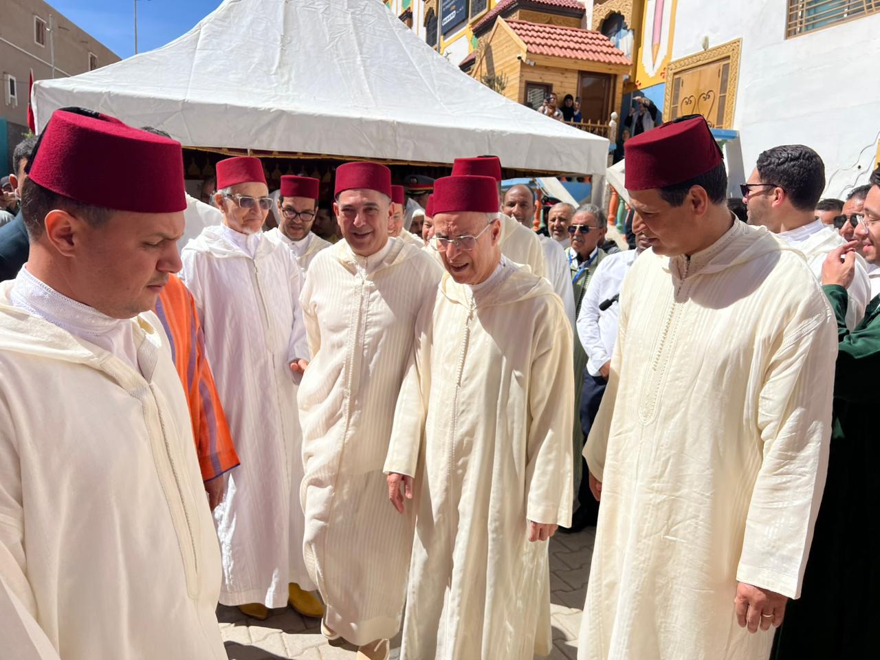 Group of men in white traditional robes and red fezzes at an outdoor ceremony near a white tent.