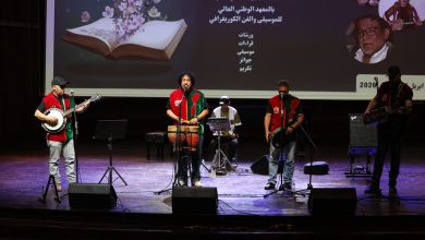A group of musicians performing on a theater stage, wearing red-and-green vests, with drums and guitars and a large screen behind them displaying Arabic text and images.