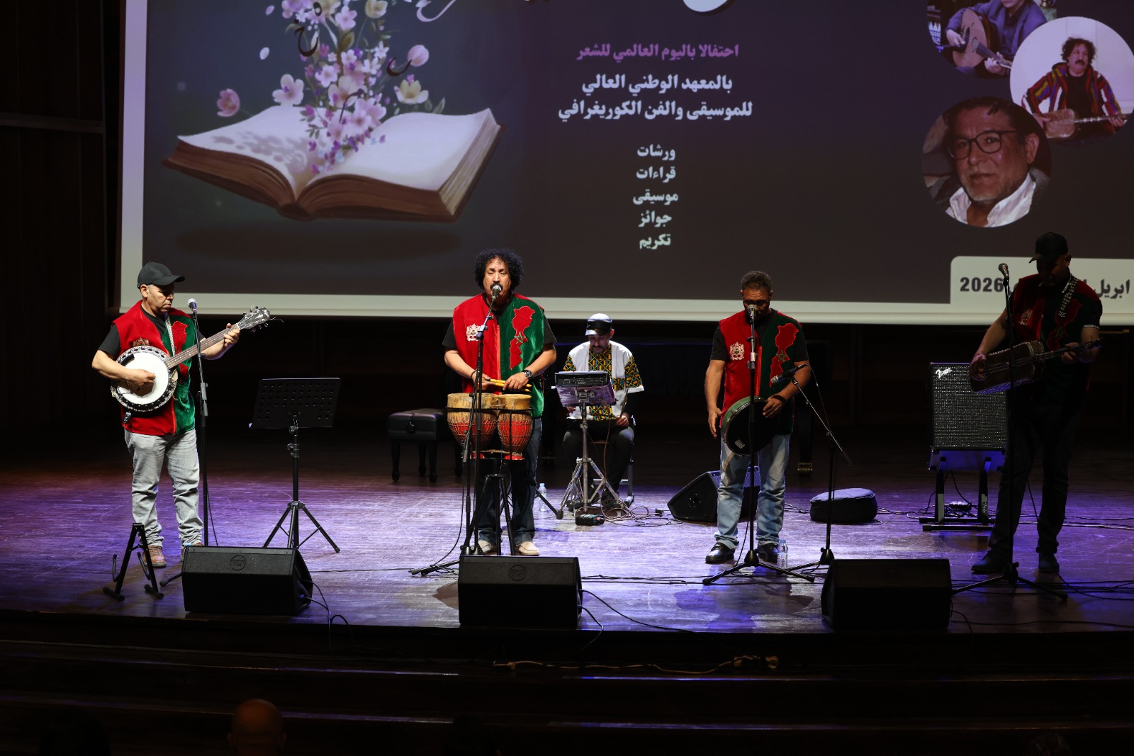 A group of musicians performing on a theater stage, wearing red-and-green vests, with drums and guitars and a large screen behind them displaying Arabic text and images.