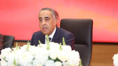 Middle-aged man in a dark suit speaks at a conference table, with white flowers in front and a red backdrop.