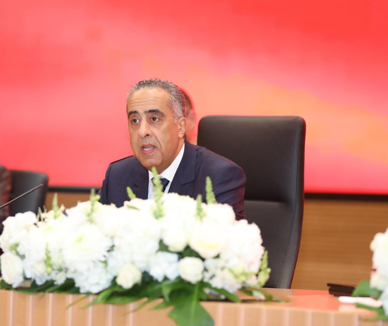 Middle-aged man in a dark suit speaks at a conference table, with white flowers in front and a red backdrop.