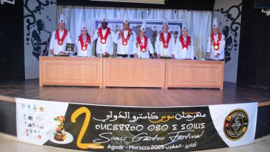 Group of chefs in white uniforms with red sashes and large medals posing behind long wooden desks on a stage with a festival banner in view."