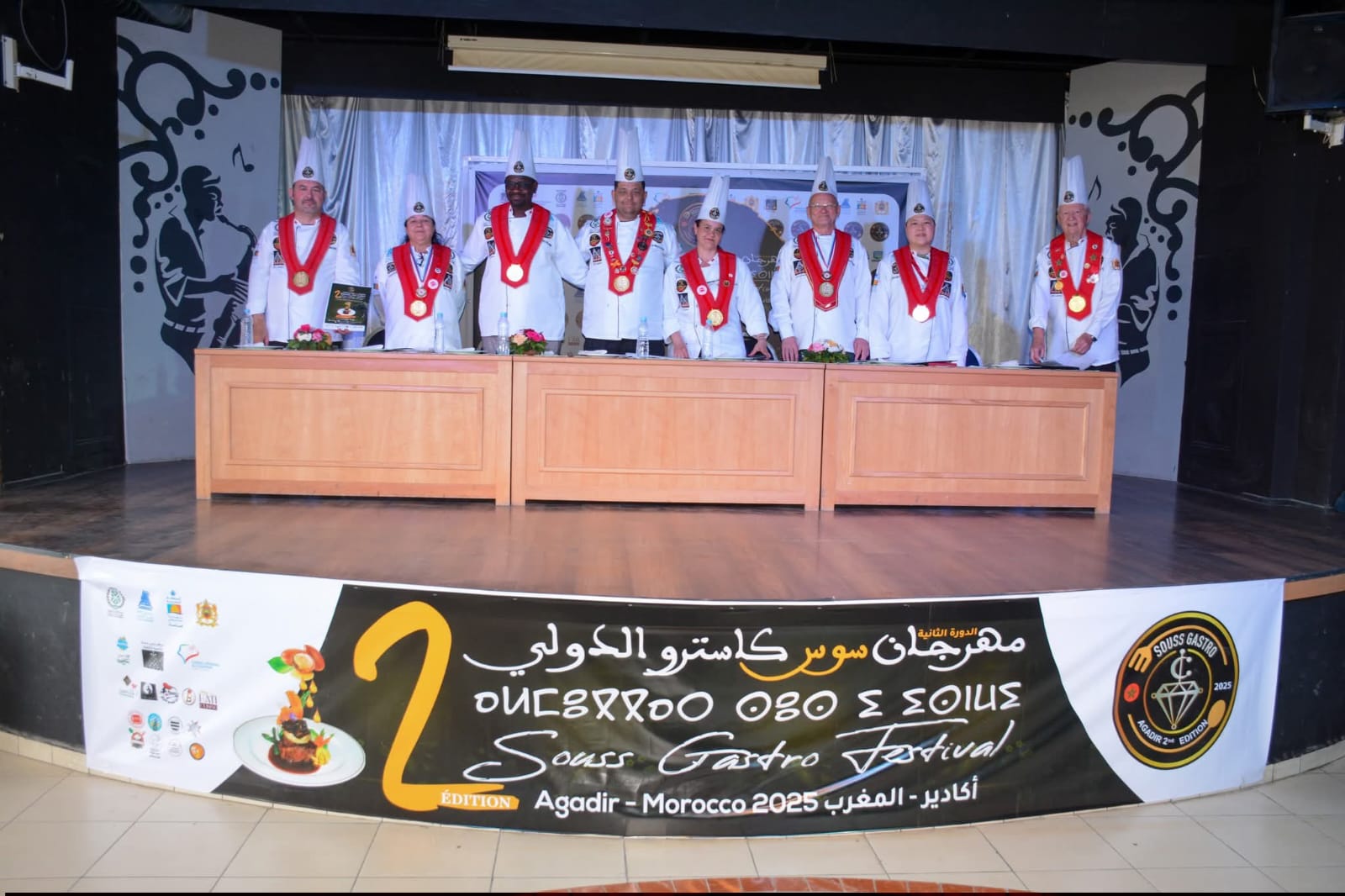 Group of chefs in white uniforms with red sashes and large medals posing behind long wooden desks on a stage with a festival banner in view."