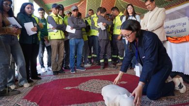 Woman kneels to perform CPR on a dummy while a group of trainees watches inside a tent with patterned fabric.